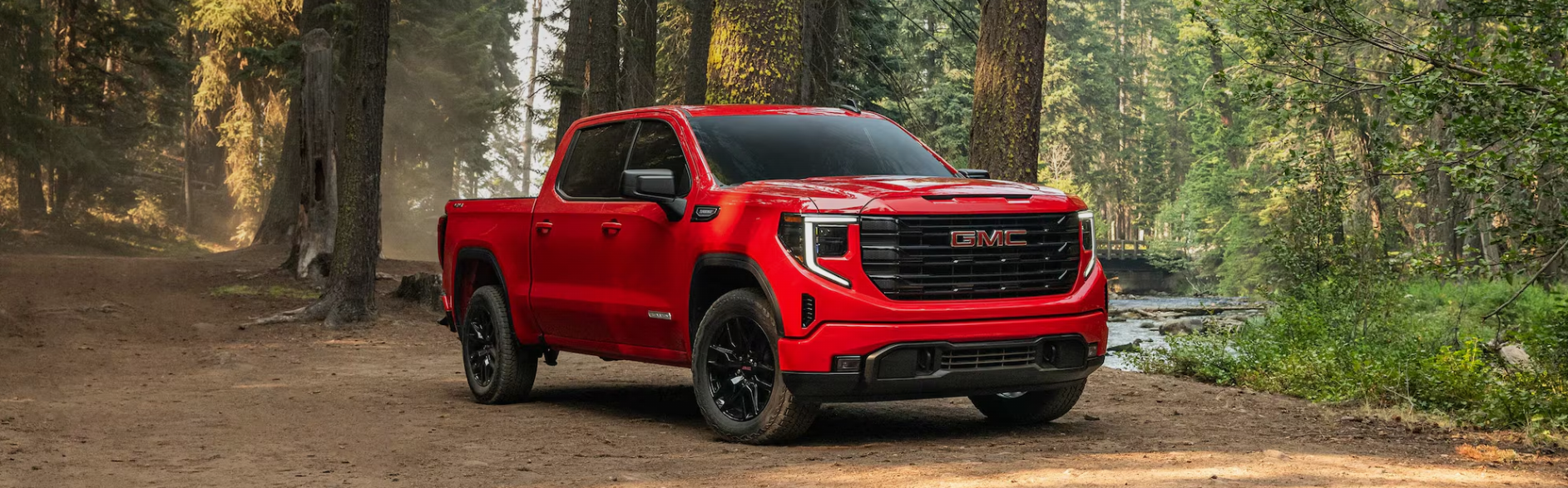 Red GMC Sierra pickup truck parked on a dirt trail in a forest near a river.