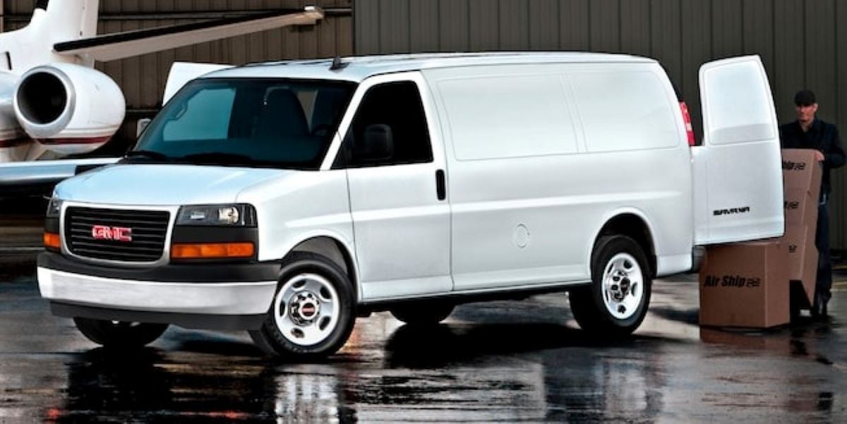 2025 GMC Savana Cargo Van parked near an airplane hangar with rear doors open and a man loading boxes.